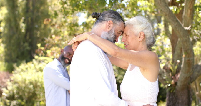 Embracing at outdoor wedding, senior bride and groom smiling with friend standing in background