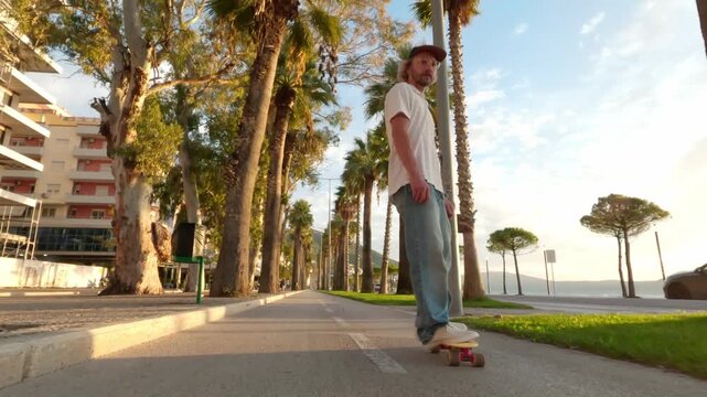 Medium shot of a young man riding while seated low on a skateboard along a palm-lined seaside promenade at golden hour. Warm sunset light, coastal city atmosphere and smooth motion create a stylish ur