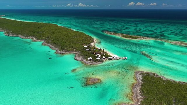 Ship Channel Cay, Exuma, Bahamas: Aerial view of a green cay tip with white cottages, a wooden dock and moored boats, descending toward turquoise tidal flats and reef channels.