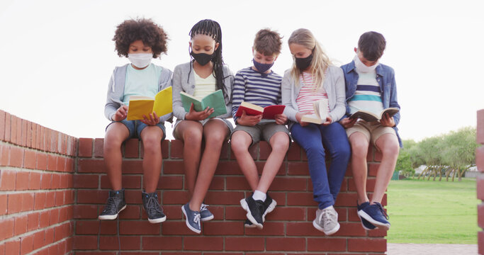 Happy diverse children in facemask sitting on wall and reading books