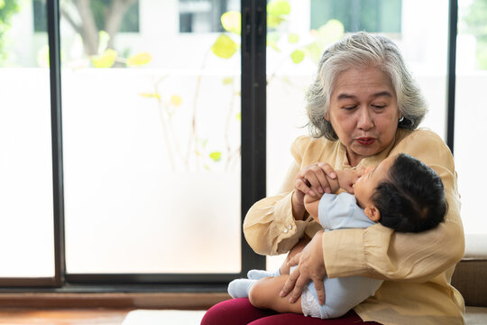 Asian grandmother tenderly holds her infant grandchild near a window at home, sharing a joyful caregiving moment for family connection, parenting, and multigenerational lifestyle campaigns