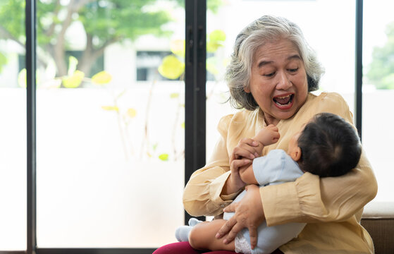 Happy Asian grandmother plays gently with her infant grandchild beside a bright window at home, showing affectionate caregiving and family connection for parenting and lifestyle advertising