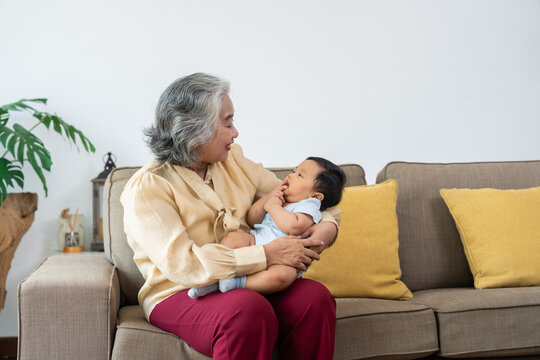 Asian grandmother gazes lovingly at her infant grandchild in her arms on a living room sofa, capturing an intimate moment for family, childcare, and senior lifestyle campaigns