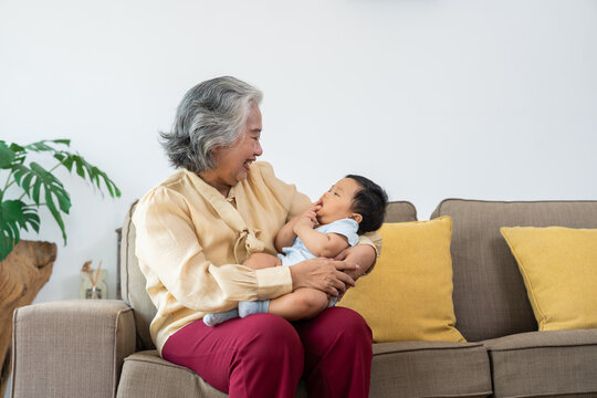 Asian grandmother smiles while holding her baby grandchild on a sofa, with a bright living room and warm yellow accents, ideal for family love, caregiving, and connection themes