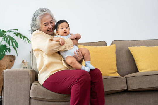 Smiling Asian grandmother cuddles her infant grandchild on a sofa in a bright living room, expressing warmth and connection for parenting, caregiving, and family lifestyle content