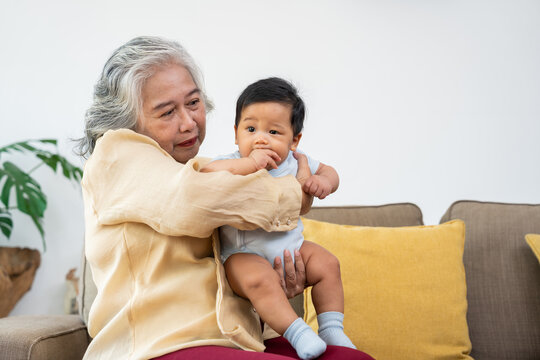 Asian grandmother holds her baby grandchild upright on her lap in a cozy living room, looking at the infant with affection, suited to family care, bonding, and healthy aging campaigns