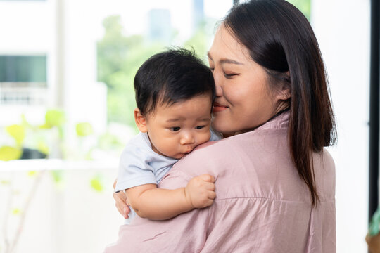 Asian mother cuddles her baby close and kisses the infant's cheek while holding the child securely at home, expressing tenderness, emotional connection, and the warmth of family life.