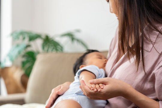 Close-up of an Asian mother supporting her resting baby and gently holding the infant's hand on a sofa, emphasizing touch, trust, and the intimate bond of everyday parenting at home
