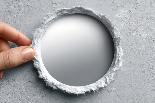 Hand touching a circular hole torn in gray paper revealing white space with background