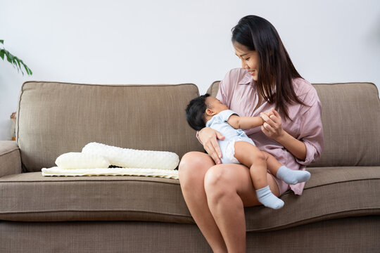 Asian mother gently cradles her sleeping baby on a living room sofa, sharing a quiet bonding moment at home with soft natural light and copy space for family care and parenting campaigns.