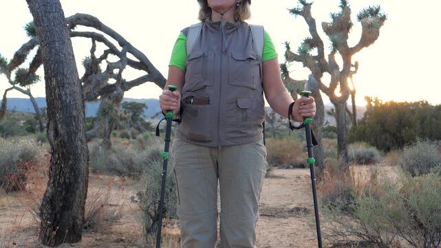 Mature female hiker holding trekking poles while standing in desert landscape of joshua tree national park during sunset. Tourist enjoying active retirement outdoor adventure. Shot from bottom to top