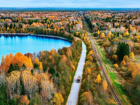 Aerial view of logging truck on lakeside road by railway tracks in autumn forest logistics