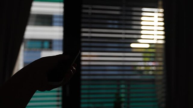 Hand holds a remote control to operate motorized window blinds in a modern apartment. Smart home automation allows convenient adjustment of daylight, privacy and indoor comfort