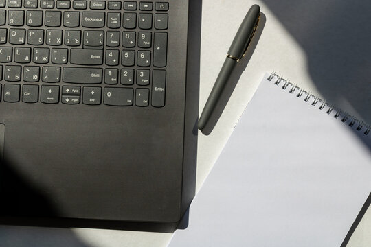 Top view of a black laptop with Cyrillic keyboard, pen, and blank spiral notepad on a white desk with sunlit shadows. Business mockup concept.