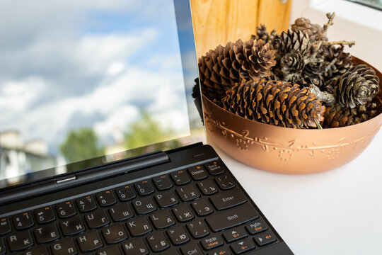 Laptop screen reflecting blue sky and clouds next to a decorative copper bowl filled with pinecones on a bright windowsill.