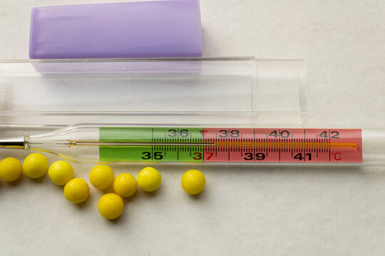 Glass medical thermometer indicating fever at 37.4C beside yellow ascorbic acid vitamin dragees on a white surface. Cold treatment concept.