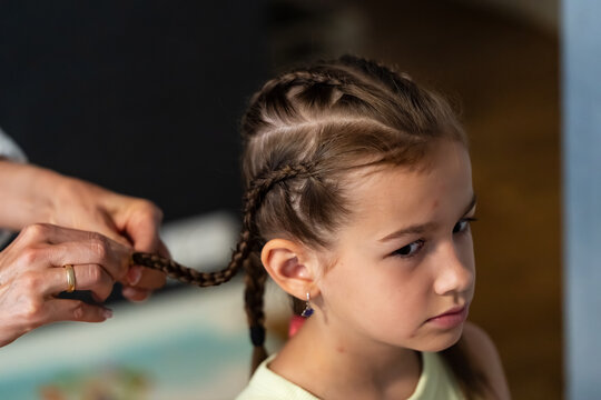 A young girl with neatly sectioned hair sits still while an adult braids her hair at home, creating intricate cornrows with warm indoor lighting and a close-up view, ideal for family lifestyle