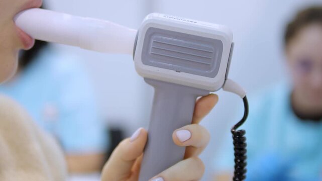 Close-up of a person using a medical device near their mouth. Hand holds gray and white instrument with coiled cable. Background shows blurred medical staff. Focus remains on device