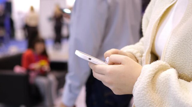 Woman holds smartphone, taps screen. Background blurred with people. Outdoors or indoor public space. Focused on hands and phone interaction