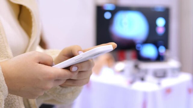 Close-up of hands holding a white smartphone. Background shows blurred tech display screens. Person wears light-colored sweater. Focus remains on device interaction