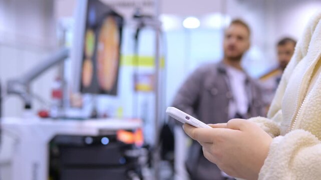 Person holds phone, focused on screen. Background shows blurred medical equipment and people. Scene appears in clinical or exhibition setting. Person wears light-colored robe or jacket