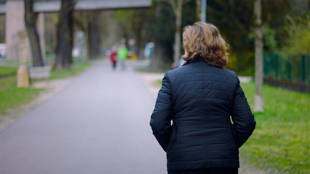 Elderly Caucasian woman walking away along a park path on a spring day, solitude and an active daily routine