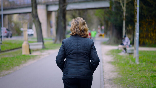 Elderly Caucasian woman walking away along a park path on a spring day, solitude and an active daily routine