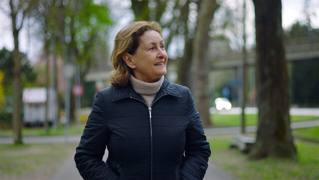Caucasian senior woman walking on a tree-lined park path with hands in pockets, peaceful stroll and wellbeing