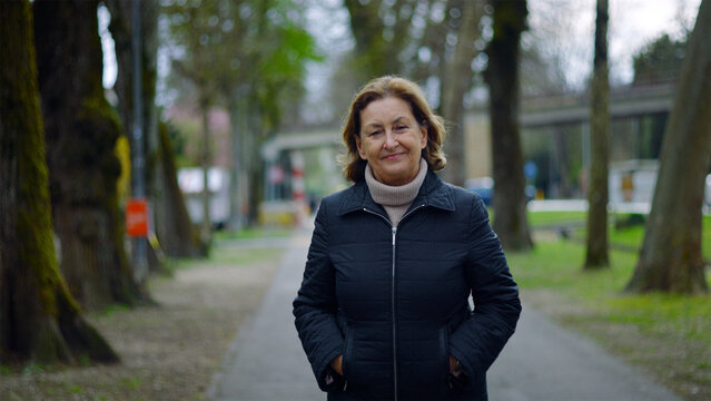 Caucasian senior woman walking on a tree-lined park path with hands in pockets, peaceful stroll and wellbeing