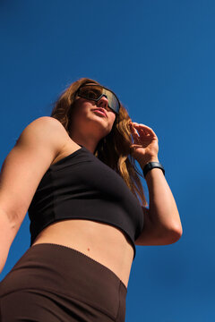 A mid-adult woman stands with one hand near her hair against a saturated blue summer sky, photographed in a vertical low