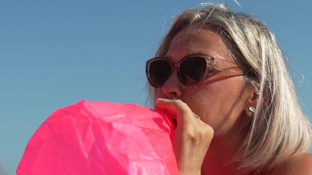 Blonde woman inflating bright pink mattress float on sunny day at seaside. Medium static shot.