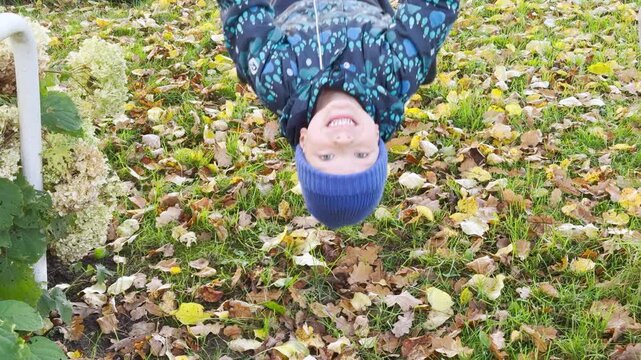 A cheerful boy hangs upside down smiling happily over fallen leaves in a park. He feels joyful, playful, and carefree during a crisp autumn day.