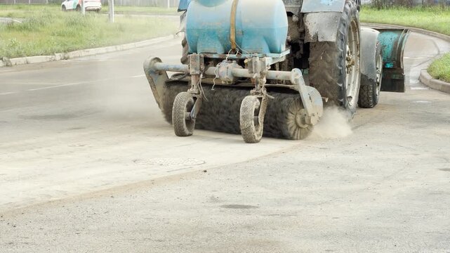 A street sweeper tractor cleans dusty asphalt with a rotating brush attachment on a sunny city road. The active scene feels productive, energetic, and satisfying.