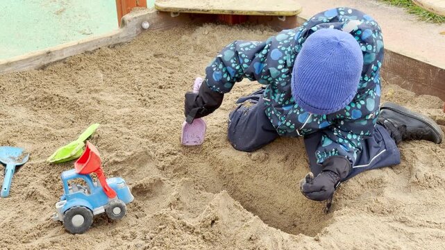 A young boy in a blue beanie plays happily in a sandbox with toy shovels and a plastic tractor. He looks focused and content on a bright outdoor playground.