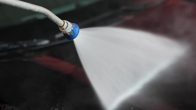 Pressure washer sprays water onto a car hood at a self service car wash. Vehicle cleaning process removes dirt from the painted surface. Water jet covers the exterior panel. 