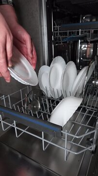 A man takes a white ceramic plate from a built-in dishwasher loaded with clean dishes and cookware