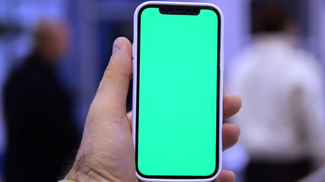 Hand holds phone with green screen. Background shows blurred people in indoor setting. Focus remains on phone throughout footage
