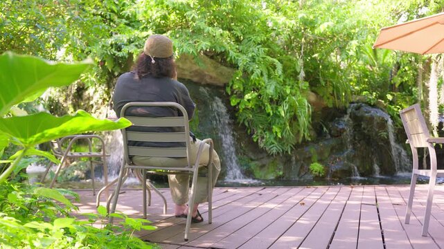 A person from behind relaxes on a patio chair, finding solitude and tranquility while contemplating a beautiful waterfall in a lush, landscaped garden oasis.