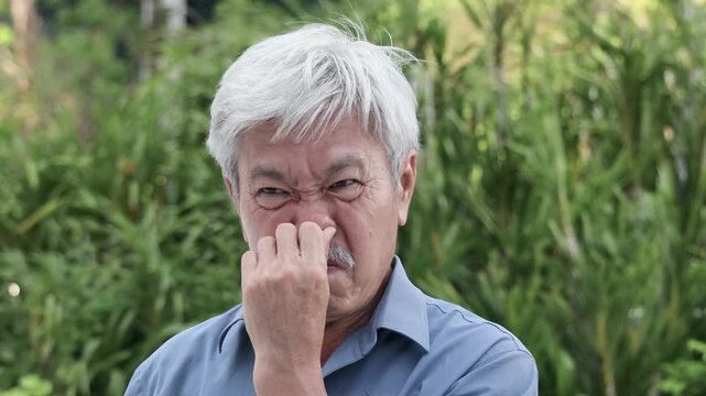 Senior Asian man covering, pinching nose to prevent bad stinking smell, stinking odor, rotten smell with disgusting expression or reaction, outdoor scene