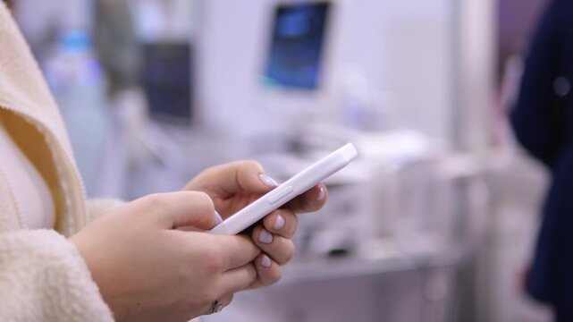 Person holds white smartphone in hands. Fingers tap screen while standing indoors. Background shows blurred tech equipment. Out of focus people nearby. Modern device use in public space