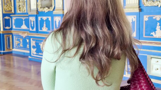 A woman admires ornate blue walls with golden details inside the Catherine Palace in Pushkin, Russia. She feels wonder and calm in the grand, elegant hall.