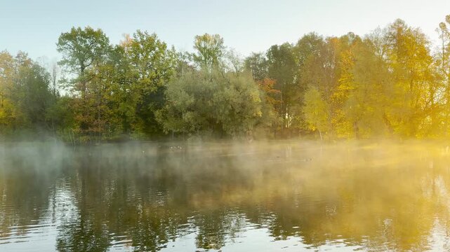 A pan shot sweeps across morning mist rising above a park lake in Pushkin in slow motion. Golden sunlight hits colorful autumn trees, bringing a warm, serene feeling.