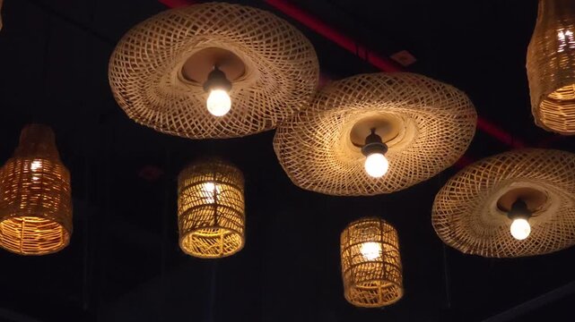 Woven bamboo basket and hat lamps hanging from indoor ceiling with glowing lightbulbs