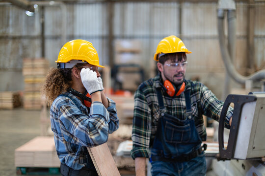 A woman in a carpentry workshop adjusts safety glasses, holding wood while her male colleague examines the machine control panel.