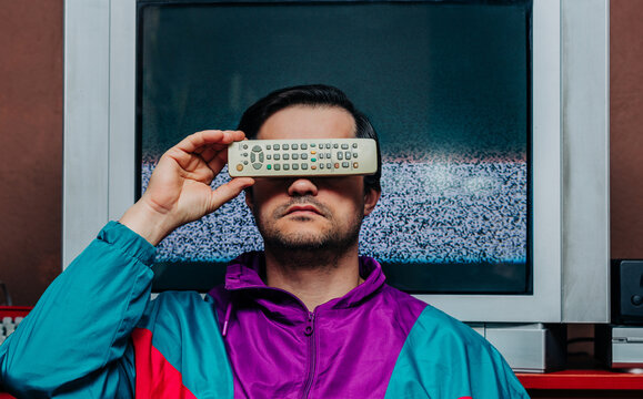 Man in 80s outfit holding TV remote over eye, static screen behind