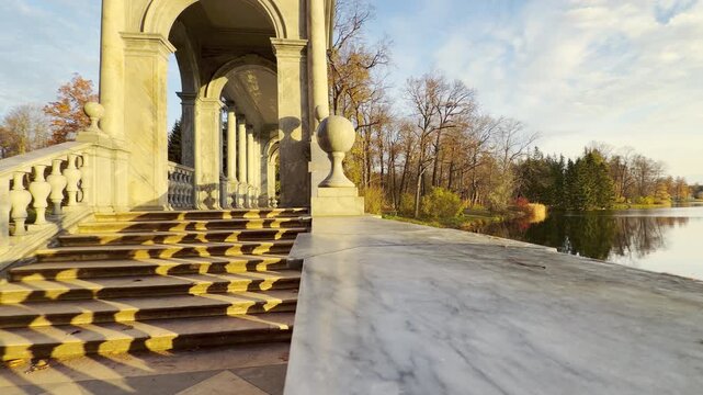 Golden sunlight casts long balustrade shadows across marble stairs leading to a classic gallery. A peaceful autumn scene in Catherine Park, Pushkin, Russia brings warm tranquil joy.