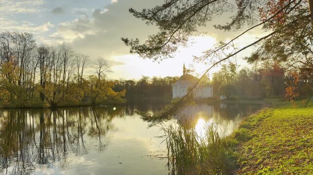 Pine branches frame the Turkish Bath pavilion as sunset reflects on calm lake waters. A peaceful autumn scene in Catherine Park, Pushkin, Russia brings warm tranquil joy.