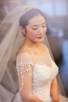 Beautiful Asian bride in white beaded wedding gown with tiara and veil, soft window light, wedding preparation portrait