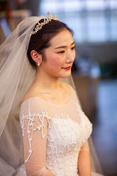 Beautiful Asian bride in white beaded wedding gown with tiara and veil, soft window light, wedding preparation portrait