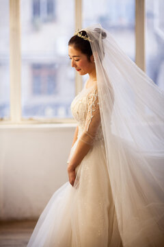 Beautiful Asian bride in white beaded wedding gown with tiara and veil, soft window light, wedding preparation portrait